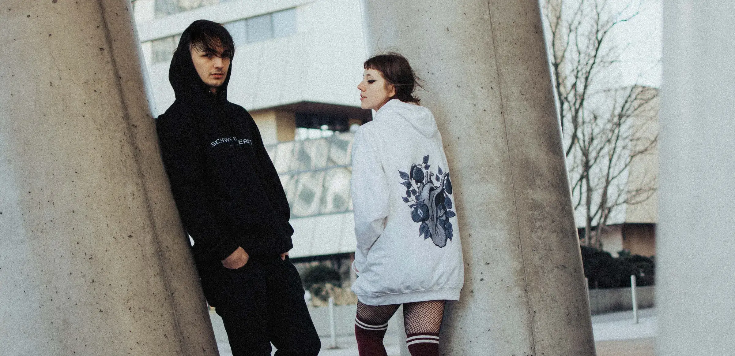 Two models in black and white Schweineheart hoodies leaning on concrete columns